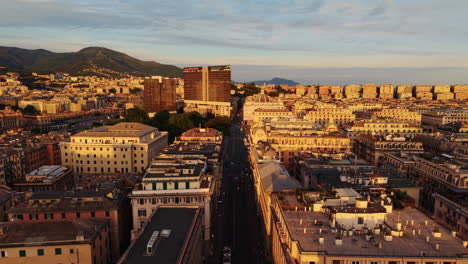 drone flies backward above a street in genoa, showing traffic lights, cars, rooftops, mix of modern and old buildings, with sea and mountains in the background