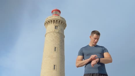 Ein-Junger-Brauner-Kaukasischer-Mann-Mit-Kurzen-Haaren,-Der-Auf-Seiner-Vernetzten-Uhr-Mit-Einem-Leuchtturm-Und-Einem-Blauen-Himmel-Im-Hintergrund-Schaut-Und-Scrollt