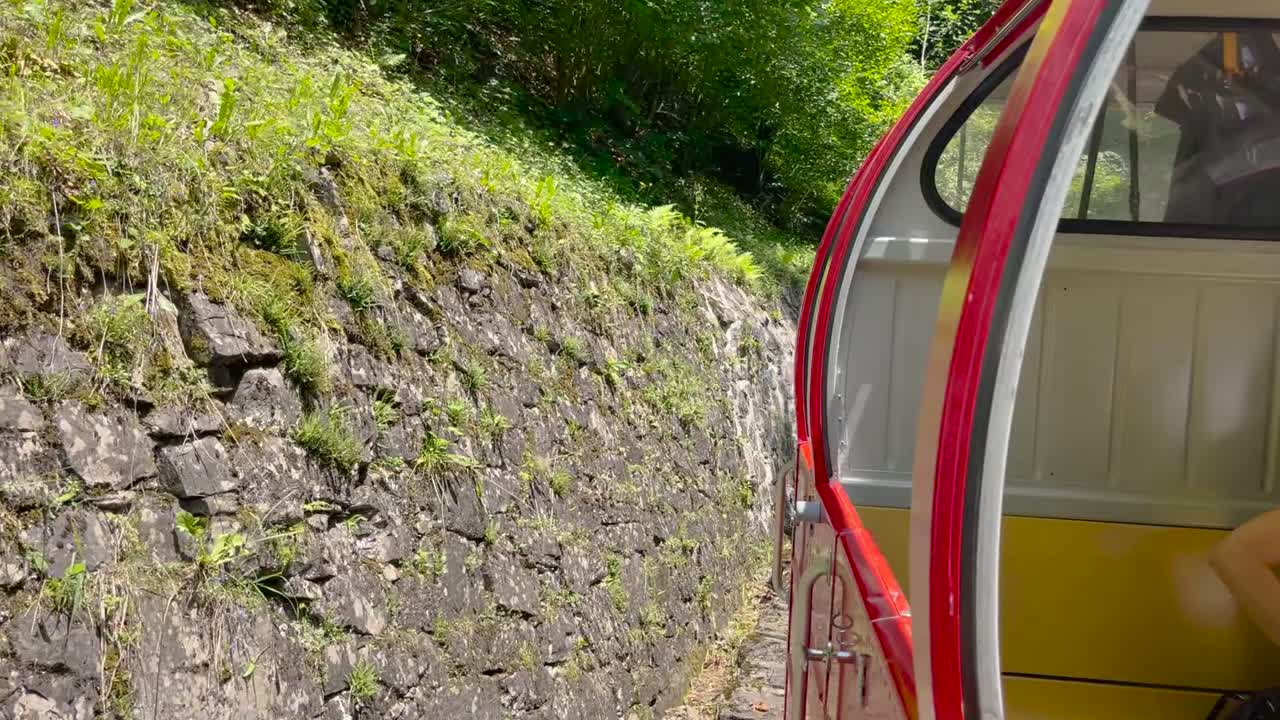 Tomada em acompanhamento ao lado de um trem a vapor vermelho e amarelo passando por uma encosta rochosa íngreme e uma floresta verde exuberante na Brienz Rothorn Bahn, Suíça, em um dia ensolarado de verão