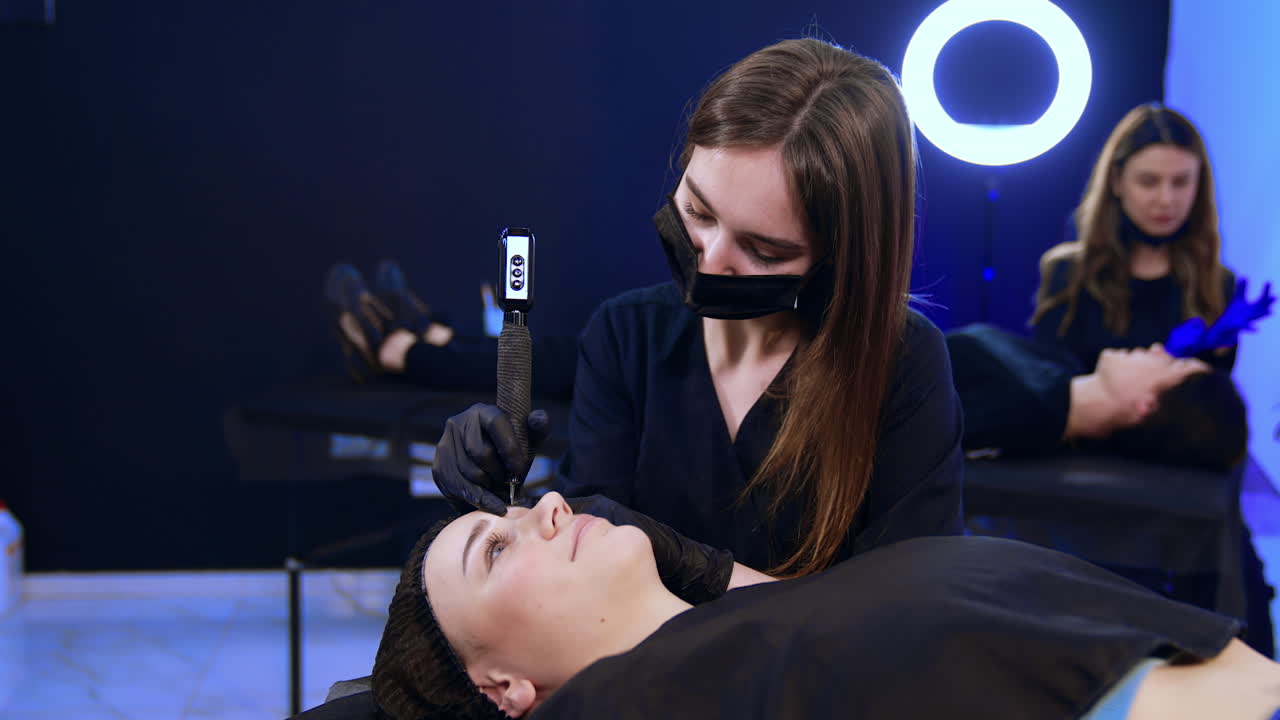 Long-haired brunette brow master uses special machine for brow make up. Specialist sits over the client lying on the couch in salon.