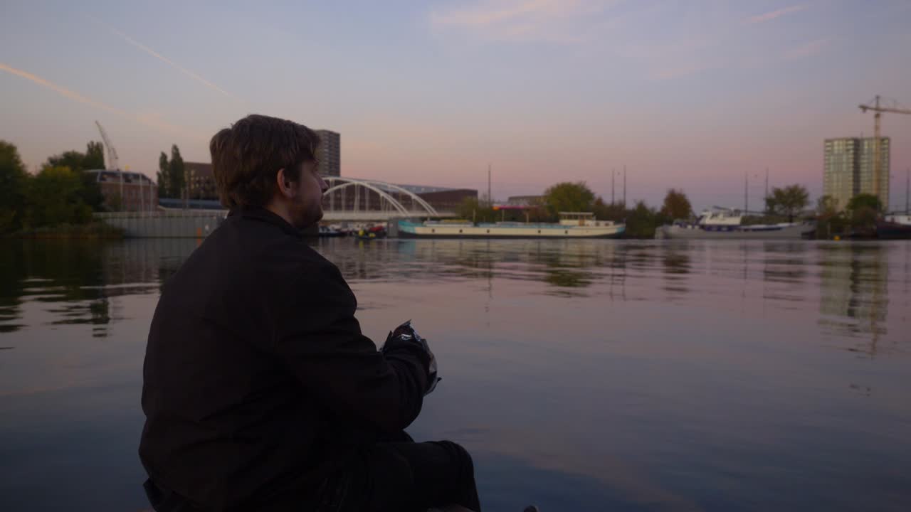 Video behind a young man sitting on the edge of a pier looking out into the water and eating potato chips during sunset