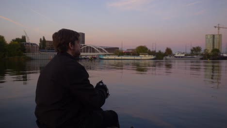 Video-behind-a-young-man-sitting-on-the-edge-of-a-pier-looking-out-into-the-water-and-eating-potato-chips-during-sunset