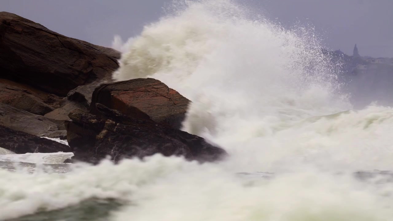 Powerful sea swell breaking violently against a large rocky outcrop with heavy white water spray. Fixed tripod telephoto shot. Overcast weather with distant coastal hills and buildings.