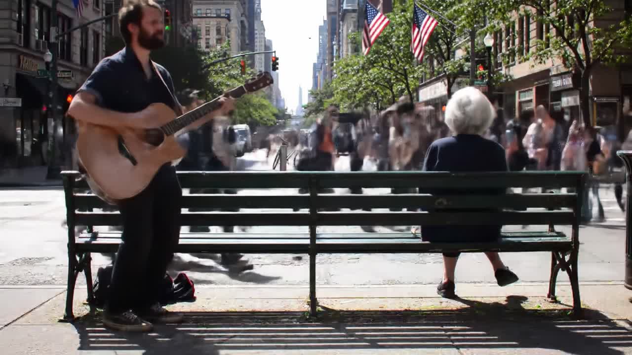 In a bustling city, a street musician captivates passersby with soothing melodies while an elderly woman sits on a bench, soaking in the vibrant atmosphere. People walk by, creating a lively backdrop.