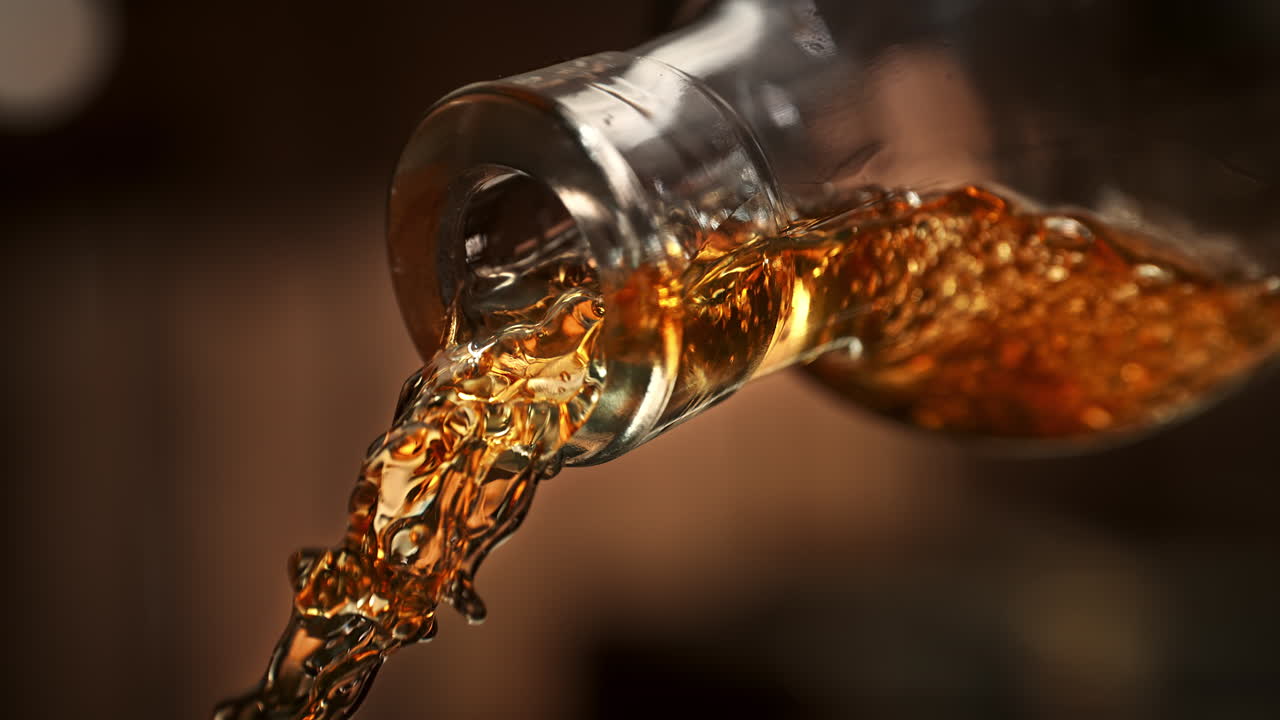 느린 동작으로 따뜻한 갈색 나무 그림자 배경에 투명한 병에서 흘러 내리는 황금색 위스키의 흐름