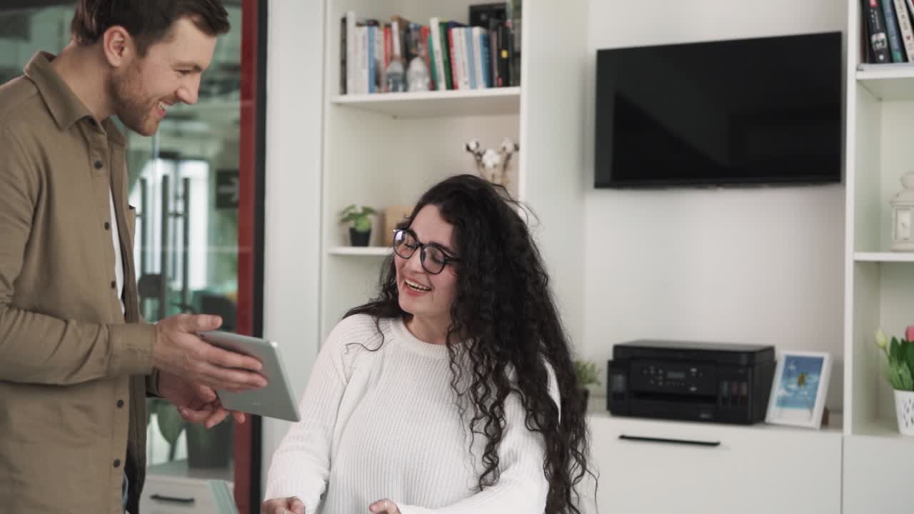 The young woman with black curly hair is sitting at a desk in the office. Her colleague approaches her and shows something on a tablet. They engage in a lively conversation