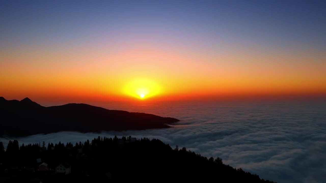 Volo sopra le montagne scure con alcune case tra gli alberi. Il sole tramonta sul paesaggio coperto da una vasta distesa di nuvole.