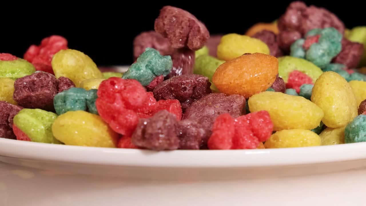 Una mano se extiende hacia un cuenco blanco lleno de vibrantes piezas de cereal multicolores con forma de fruta