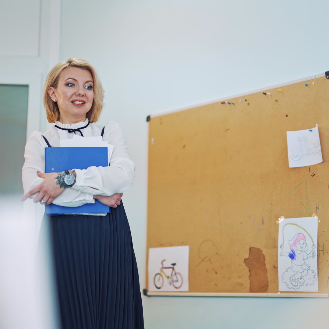 Teacher is talking in the class. Attractive teacher holding a blue folder telling something to the kids. Female educator stands near the pinboard in the classroom background.