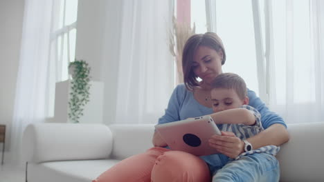 Madre-E-Hijo-Usando-Una-Tableta-Mirando-La-Pantalla-Y-Hablando-En-Un-Sofá-En-Una-Casa-Acogedora