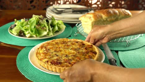 male senior hand putting a white ceramic dish of ham cheese quiche on blue table mat with a plate of various vegetable, a loaf of freshly baked bread on metal cooling rack.