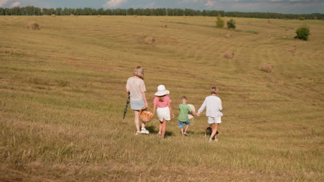 auntie and son walk across sunlit countryside carrying a hat, while mother and daughter stroll alongside with basket in hand, surrounded by rolling fields, hay bales, and distant tree