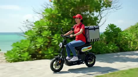 a delivery rider in red attire confidently rides an electric scooter along a scenic coastal path lined with greenery. the bright sun adds warmth to the lovely day.