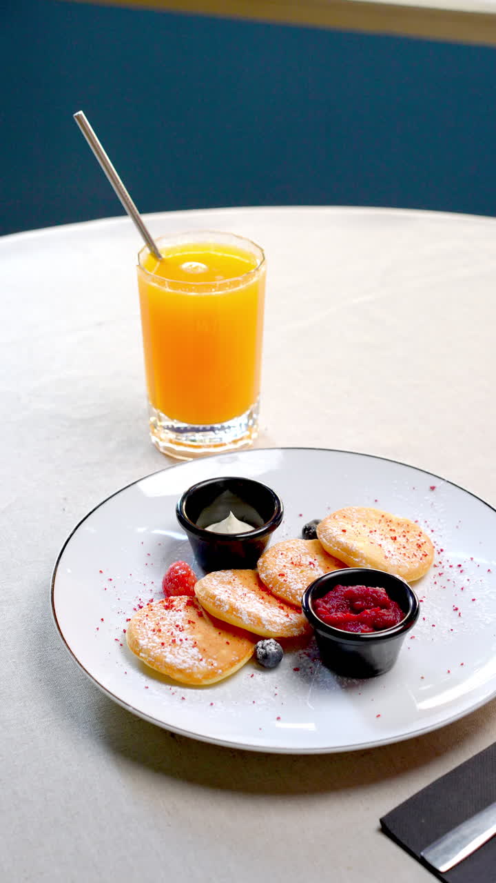 Miniature pancakes topped with vibrant forest berries, homemade jam, dolloping whipped cream alongside freshly squeezed orange juice resting on white ceramic plate near rustic wooden backdrop