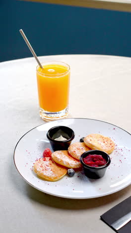 miniature pancakes topped with vibrant forest berries, homemade jam, dolloping whipped cream alongside freshly squeezed orange juice resting on white ceramic plate near rustic wooden backdrop