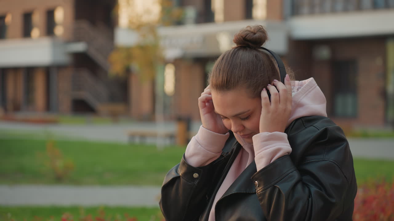 Woman in leather jacket puts on headphone enjoying peaceful music in open park setting with blurred buildings in background and vibrant green lawn surrounded by gentle morning breeze