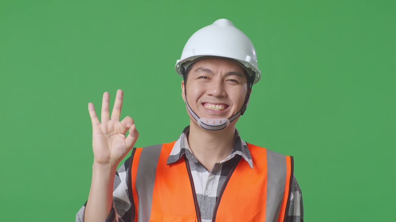 Close Up Of Asian Male Engineer With Safety Helmet Smiling And Showing Okay Gesture To The Camera While Standing In The Green Screen Background Studio