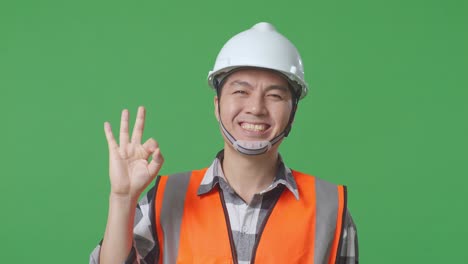 close up of asian male engineer with safety helmet smiling and showing okay gesture to the camera while standing in the green screen background studio