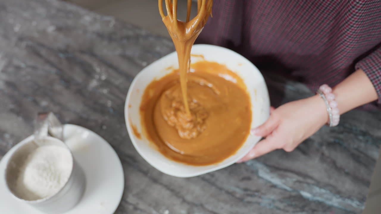 Top-down view of cook holding whisk with thick caramel-colored pap stretching as it drips back into white bowl, flour-dusted countertop and sifter nearby