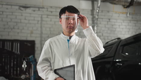 electrical engineer wearing lab coat and protective glasses adjusts eyewear while holding tablet in automotive workshop, background includes parked car, tools, and industrial workspace