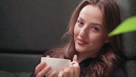 lady with long wavy hair smiles warmly at camera while holding white cup with both hands. calm indoor setting with green plant accent adds charm to cozy, relaxed coffee moment
