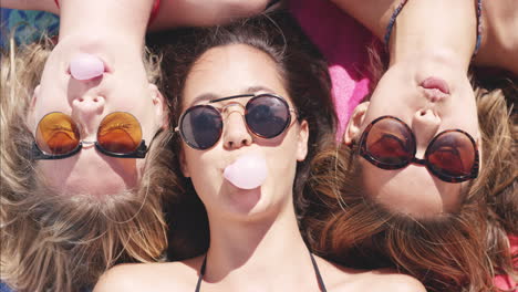 vista superior de tres amigas adolescentes acostadas de espaldas soplando burbujas de caramelo en la playa