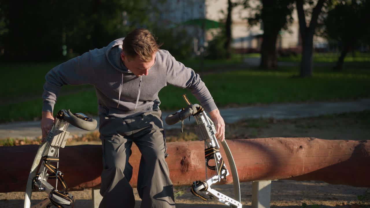 공원에서 야외에서 고 앉아있는 남자가 은 나무 줄기 위에 봄의 기둥을 들고, 회색 후디와 어두운 바지를 입고, 심각한 표정, 밝은 빛 하늘 아래 녹색 풀과 나무로 둘러싸여 있습니다.