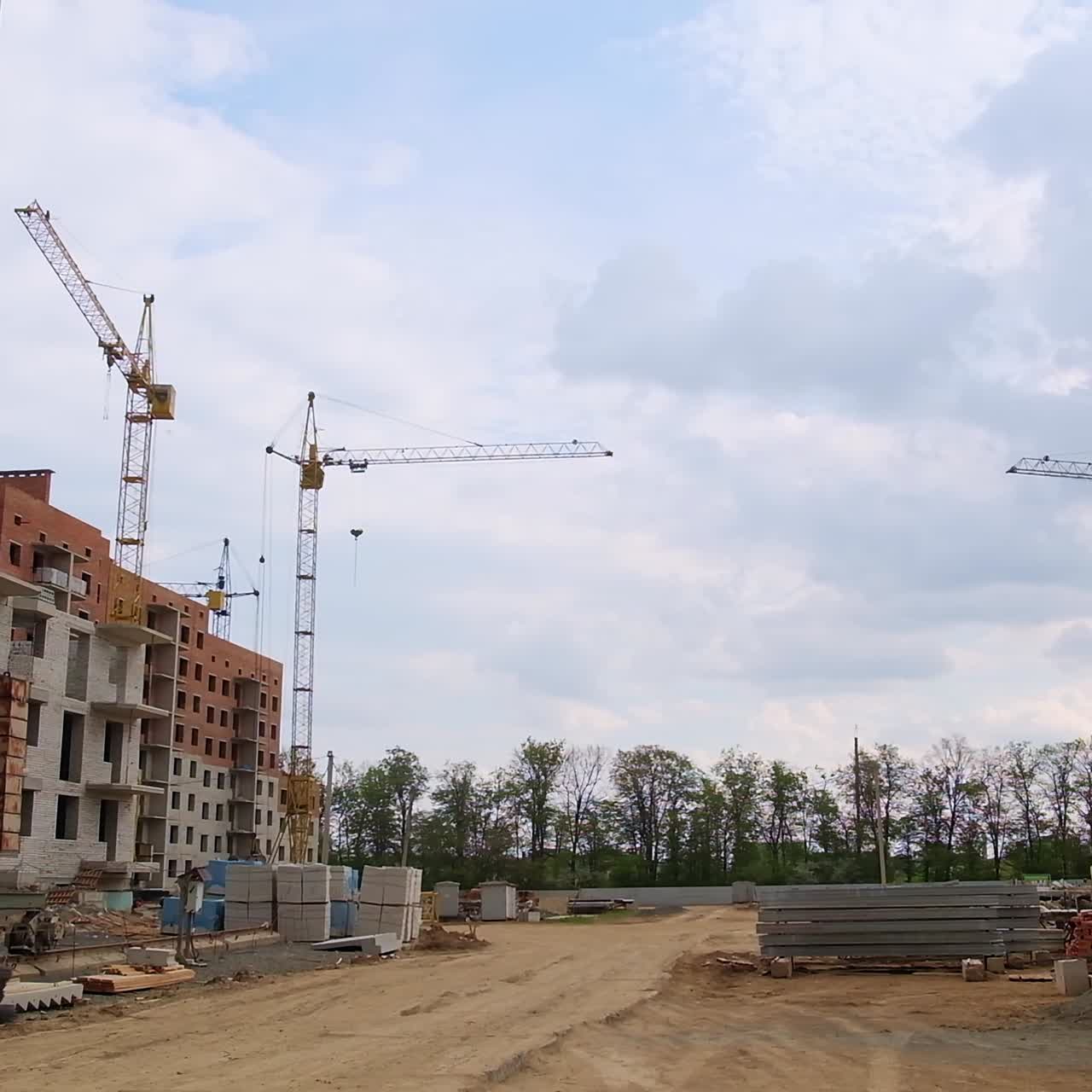 Several cranes situated on the construction site of multistoried building. Diverse building materials on the ground. Trees growing at backdrop