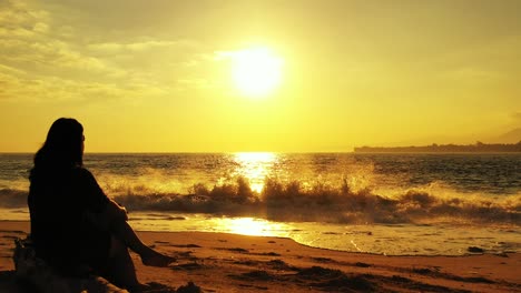 silhouette of woman sitting on quiet beach at sunset with glowing yellow sky over troubled sea with waves splashing on sand, vietnam