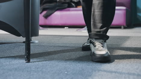 close-up of person wearing grey trousers and sneakers walking on carpeted floor. shadow and motion emphasize movement. soft lighting and modern casual style. red sofa visible in background