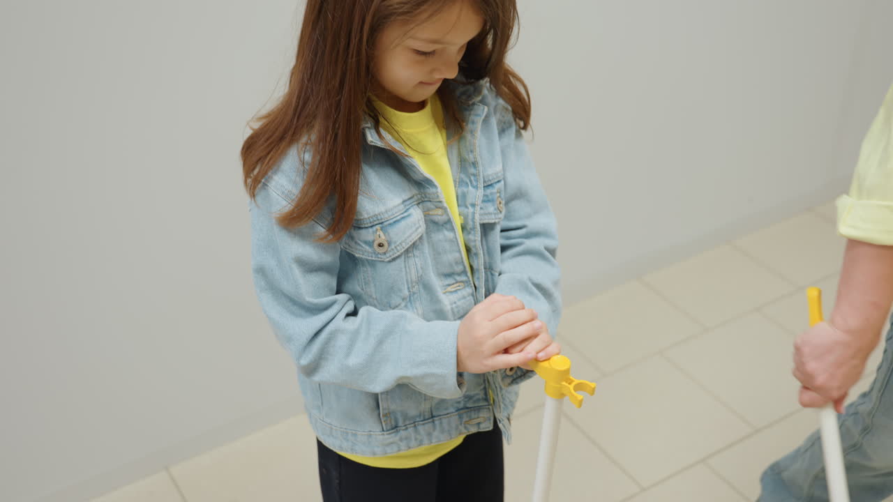 High angle view shows kid in denim holding yellow dustpan for mom while she sweeps tiled laundromat floor, packing dirt into pan, teamwork, cheerful family cleanliness scene near stainless washers