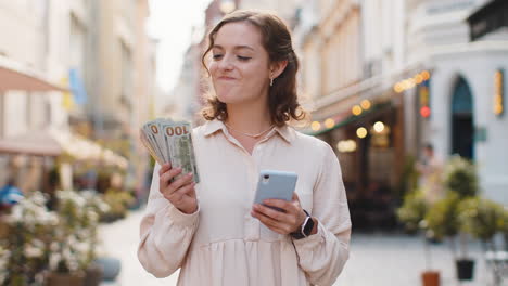 Happy-young-woman-counting-money-dollar-cash,-use-smartphone-calculator-app-in-urban-city-street