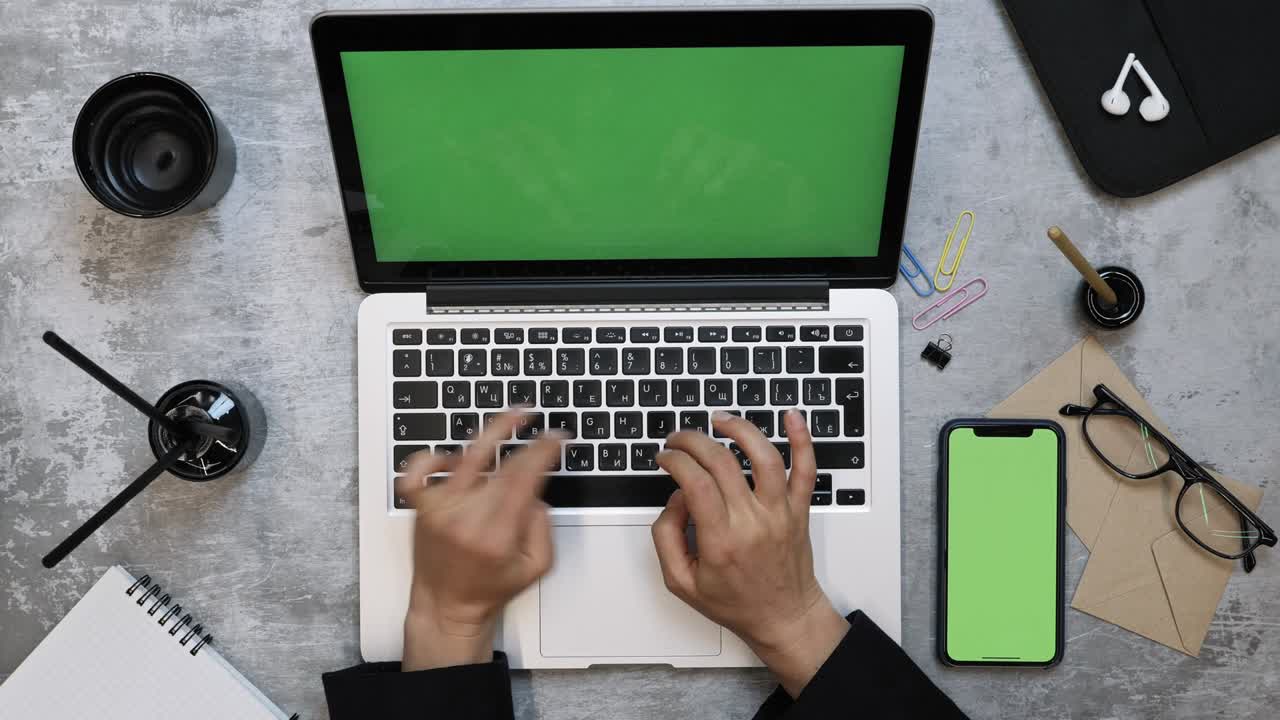Female hands working on laptop computer at office desk, view from above. Office working desk with notebook laptop and woman fingers typing on keyboard, smartphone with green screen is on right