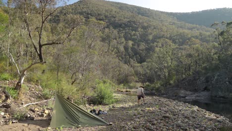 a bushman sets up a campsire with a tarp in the australian mountains by a river