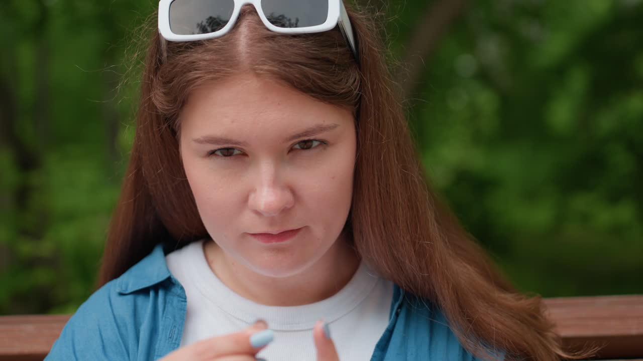 Joven sentada al aire libre en un banco del parque, concentrada y pinchándose el pulgar con una aguja, con camisa azul informal y gafas de sol blancas en la cabeza, rodeada de un suave fondo de bosque verde veraniego.