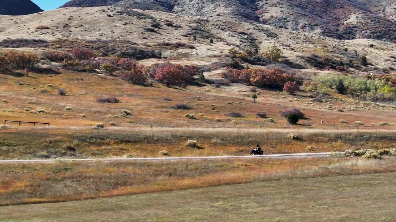 Filmisk bred luftdroneoptagelse følger en solomotorcyklist, der kører hurtigt rundt om et sving nær Deer Creek Canyon, omgivet af levende efterårsløv og Colorados forbjerge, der dominerer baggrunden.
