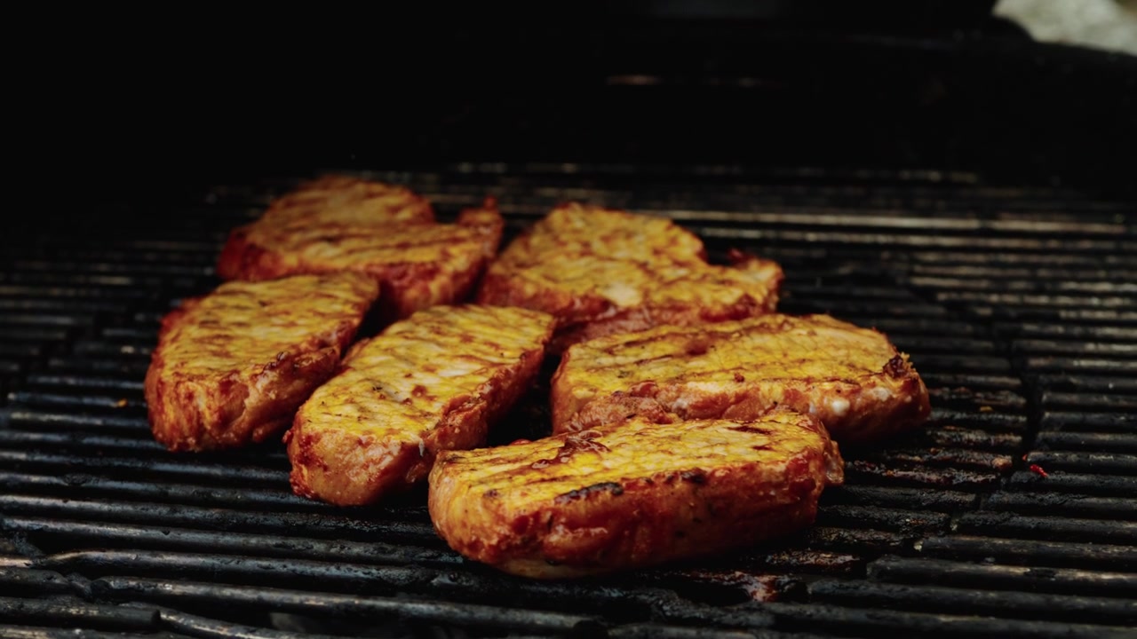 Flere krydderi-gnedne svinekødsudskæringer tilberedes sammen på en hjemmegrill med mørke grillstriber og let røg.