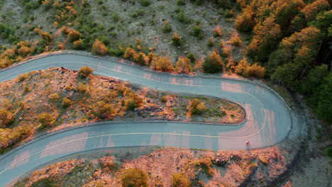 Aerial-top-view-of-a-mountain-road-with-sharp-hairpin-bend,-longboard-rider-and-car-in-shot,-golden-sunlight