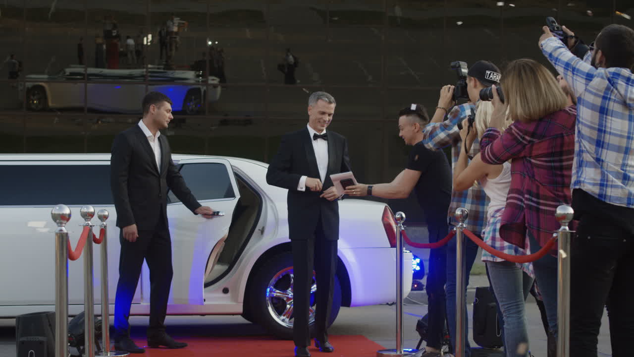 A celebrity arrives at a red carpet event, interacting with paparazzi and fans by a limousine