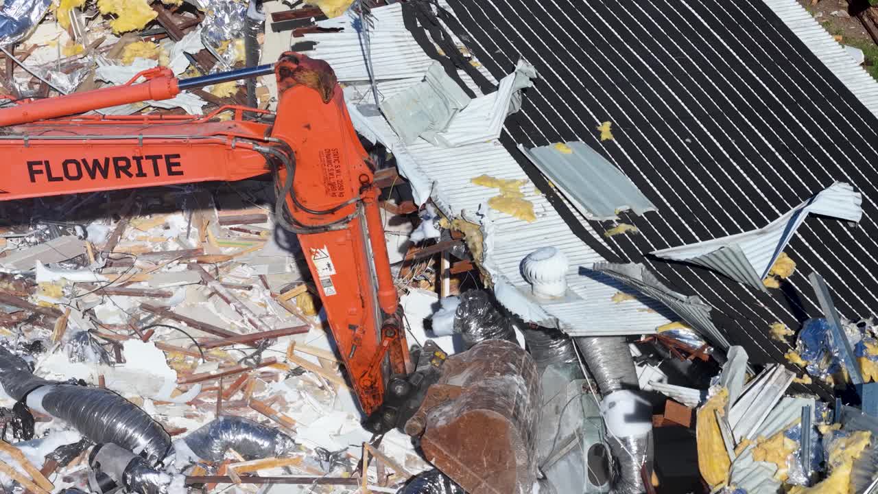 An orange excavator arm demolishes a house, moving through construction debris and scrap materials in bright daylight. Close-up aerial perspective, steady camera