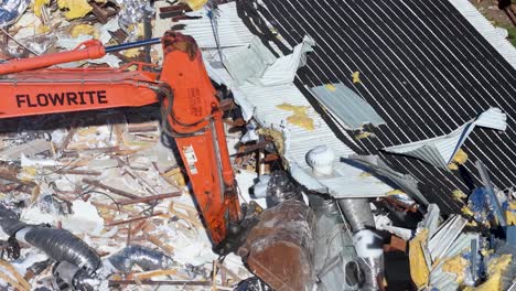 an orange excavator arm demolishes a house, moving through construction debris and scrap materials in bright daylight. close-up aerial perspective, steady camera