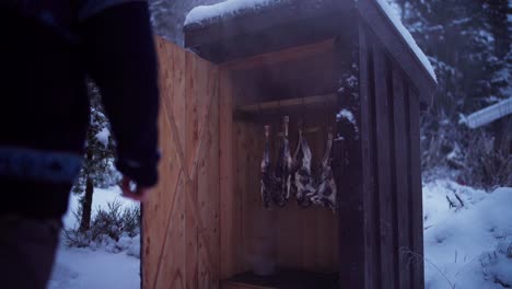 man checks on deer meat smoking inside smokehouse shed in norway during winter