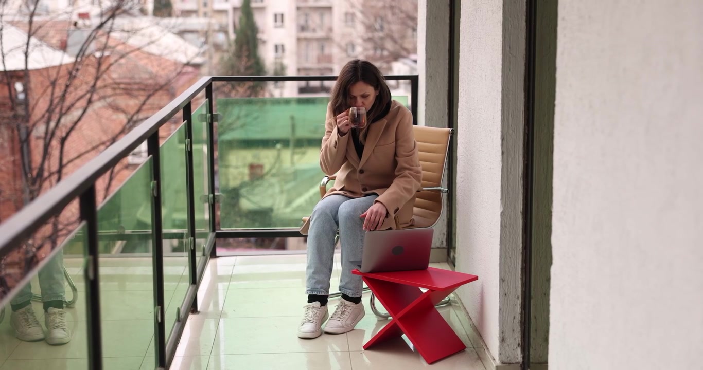 Giovane donna in cappotto lavora al laptop bevendo caffè sul balcone