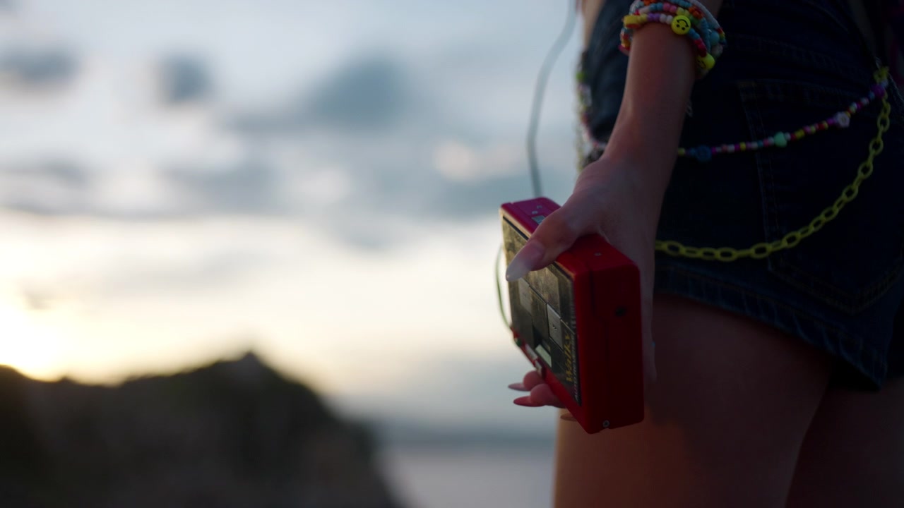 Giovane donna tiene un lettore di cassette analogico rosso portatile accanto a una baia rocciosa con la luce della sera, primo piano statico.
