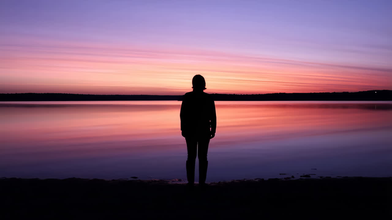 A person stands in silhouette against a vibrant sunset over a calm lake