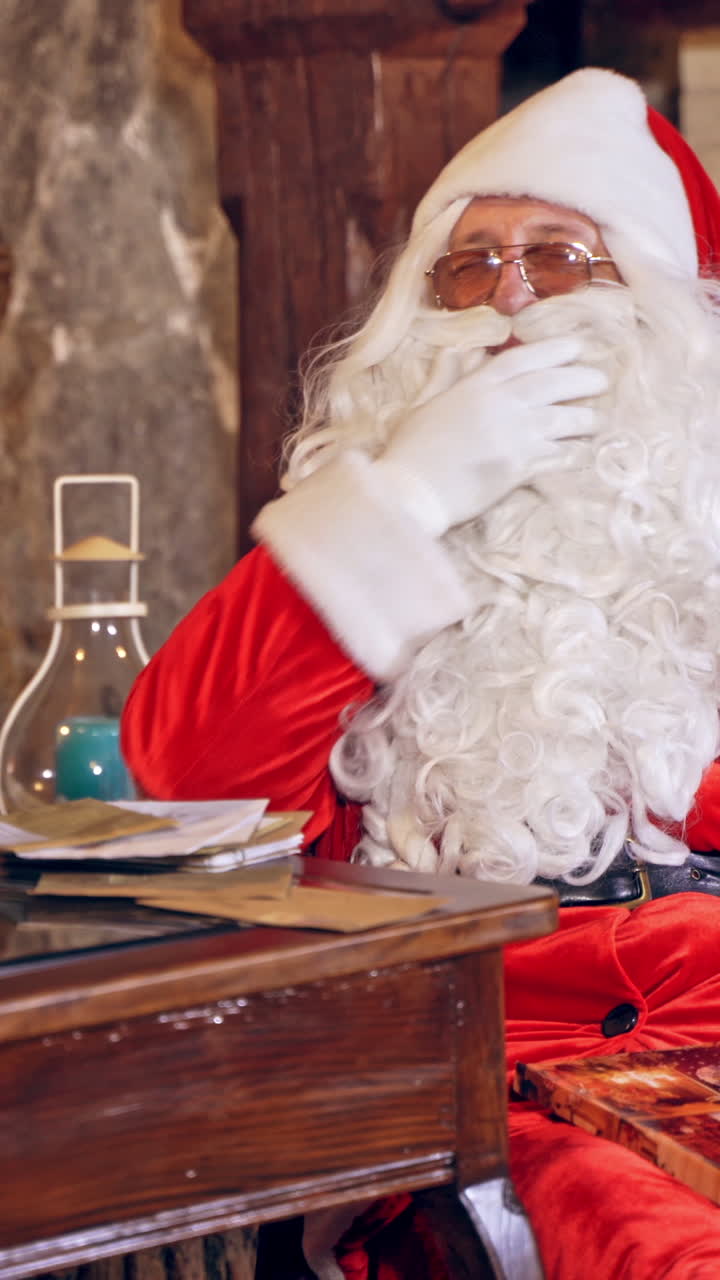 Santa Claus is sitting in specially decorated room at the table, holding an envelope in hands. Feast fireplace in the background. Vertical video