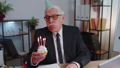 Un-Hombre-De-Negocios-Senior-Celebrando-Un-Cumpleaños-Solitario-En-La-Oficina-Soplando-Velas-En-Un-Pastel-Pequeño-Pidiendo-Un-Deseo