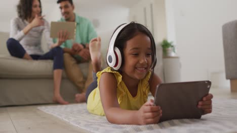 Happy-hispanic-girl-watching-video-on-tablet-with-parents-sitting-on-sofa-in-background