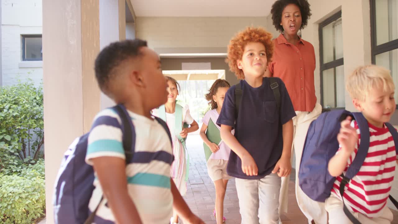 Diverse kids with backpacks exiting walkway toward yard, speeding with teacher holding tablet