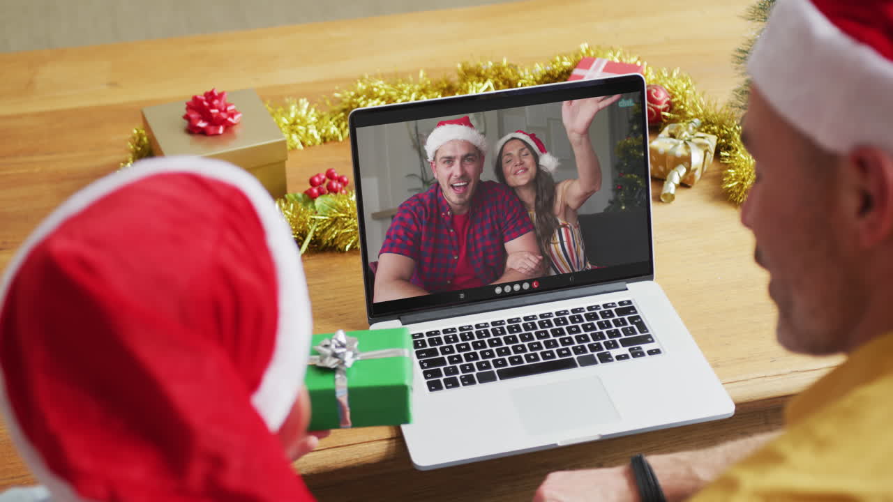 padre caucásico con su hijo usando una computadora portátil para una videollamada de navidad con una pareja sonriente en la pantalla